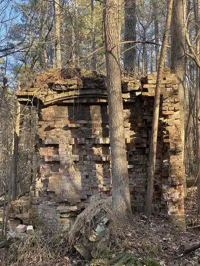 A dilapidated brick structure, possibly an old chimney or furnace, stands partially collapsed in a forest with bare trees…