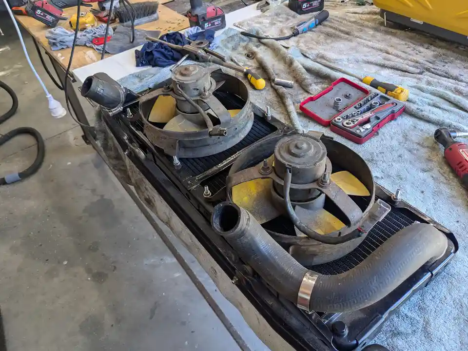 A disassembled car radiator and cooling fan assembly sits on a workbench surrounded by tools like screwdrivers, socket sets…