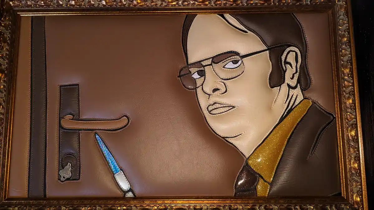 A leather art piece in an ornate gold frame depicts a stylized portrait of a man with glasses and a gold glitter tie…
