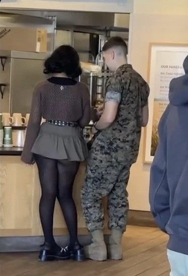 A woman in a dark skirt and tights stands at a coffee counter, facing away from the camera, while a man in a military…
