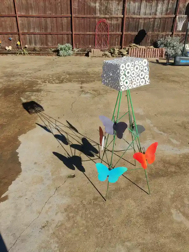 A metal garden ornament with colorful butterfly cutouts and a perforated top casts long shadows on a concrete patio, with…