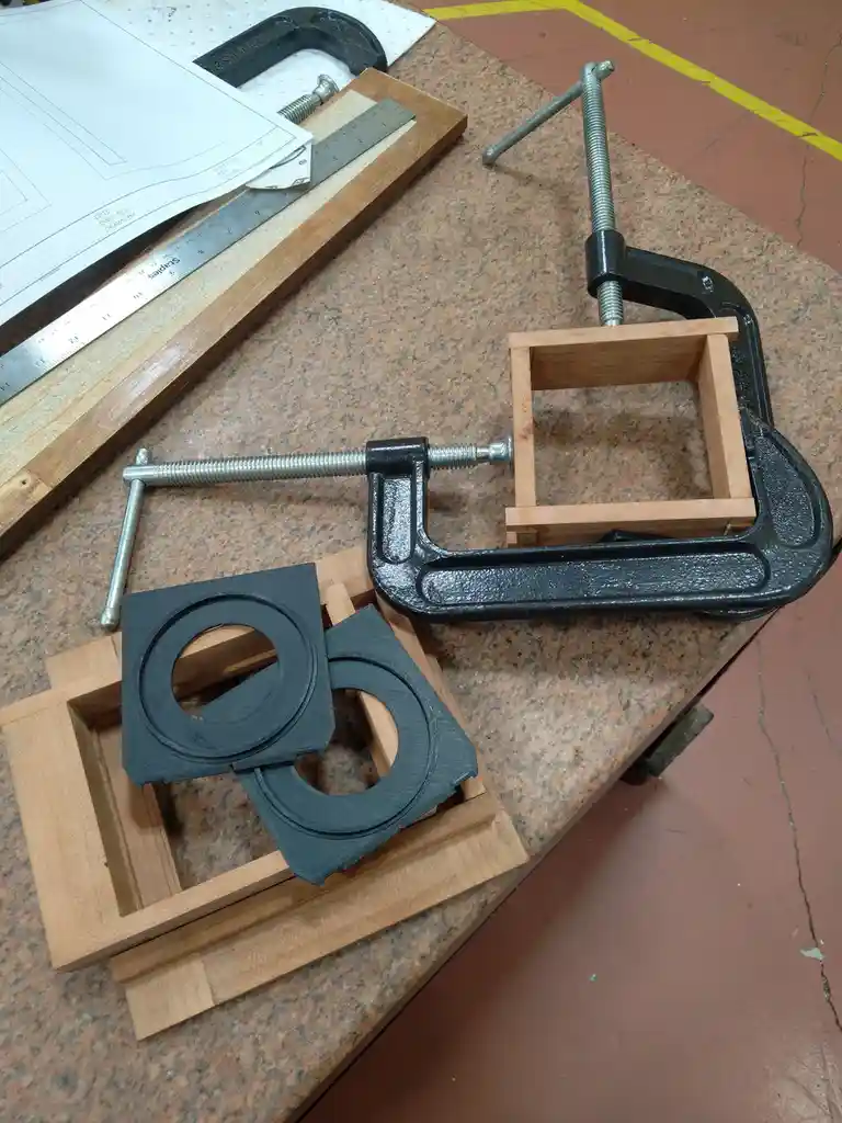 A workshop scene shows two black C-clamps holding wooden frames on a speckled workbench. One clamp holds a small square…