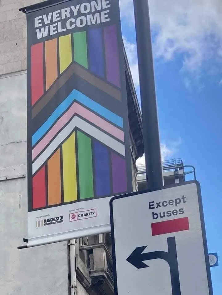 A large rainbow-patterned banner on a building reads 'EVERYONE WELCOME' with logos for Manchester Accommodation Co and a…