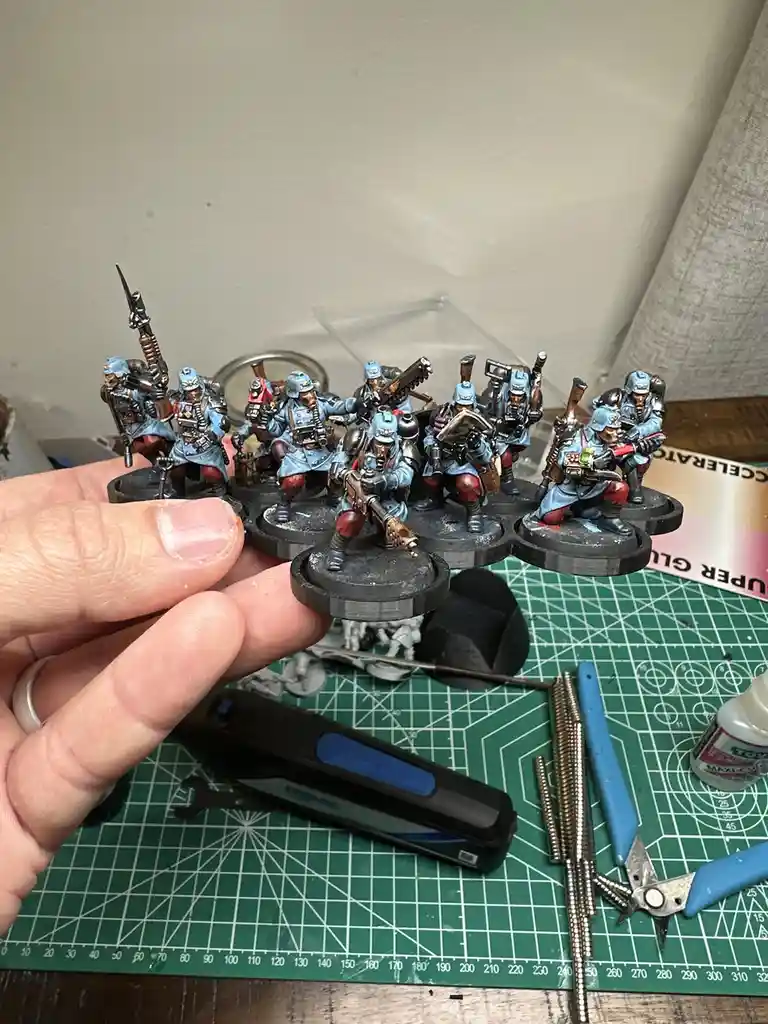 A hand holds a group of painted miniature soldiers in blue uniforms and red pants, each on a black circular base, against a…