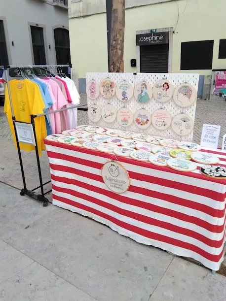 An outdoor market stall covered with a red and white striped tablecloth displays numerous hand-embroidered circular artworks…