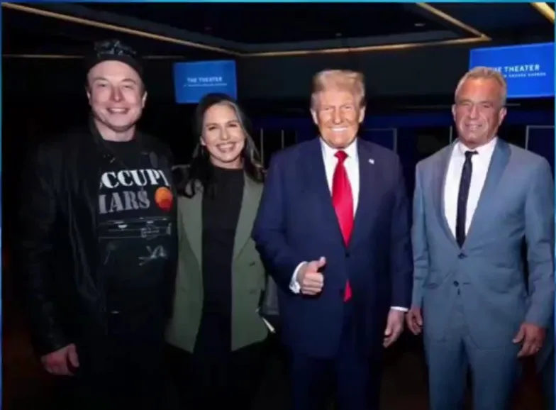 four people pose for a photo in a theater lobby. From left to right: Elon Musk in a black jacket and 'OCCUPY MARS' t-shirt…