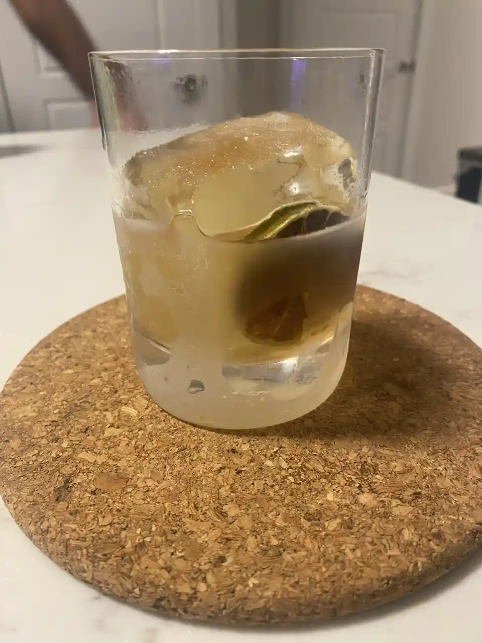 A clear glass containing a light-colored cocktail with a large ice cube and a lime wedge sits on a round cork coaster on a…