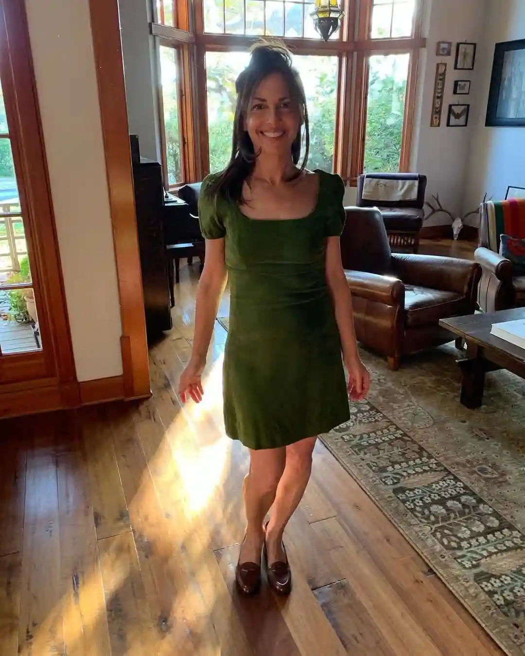 A woman with dark hair in a messy bun smiles at the camera while wearing a short-sleeved, olive-green velvet dress and brown…