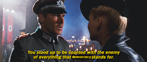 A still from a movie shows a man in a military uniform speaking to a woman in a leather jacket. The background features Nazi…
