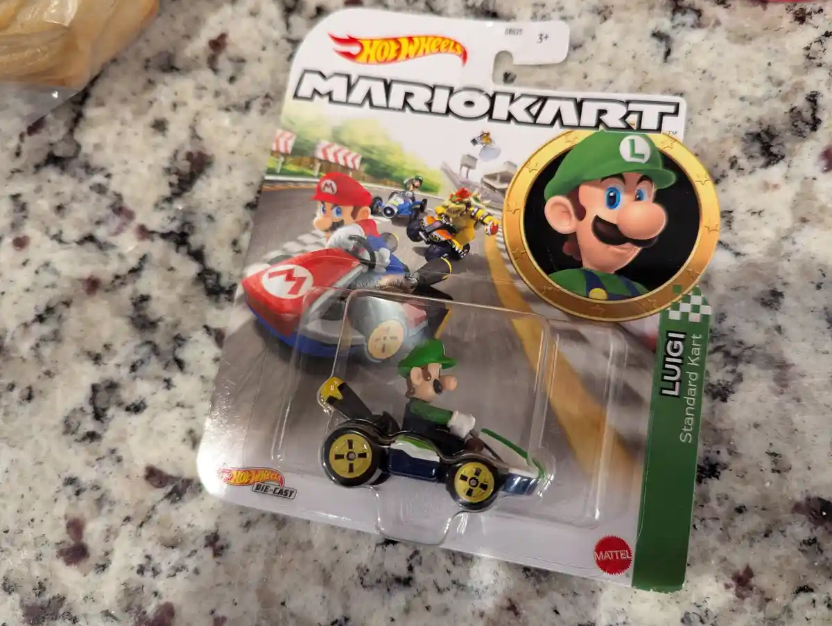 A Hot Wheels Mario Kart Luigi die-cast toy car in its original packaging sits on a speckled countertop. The packaging…