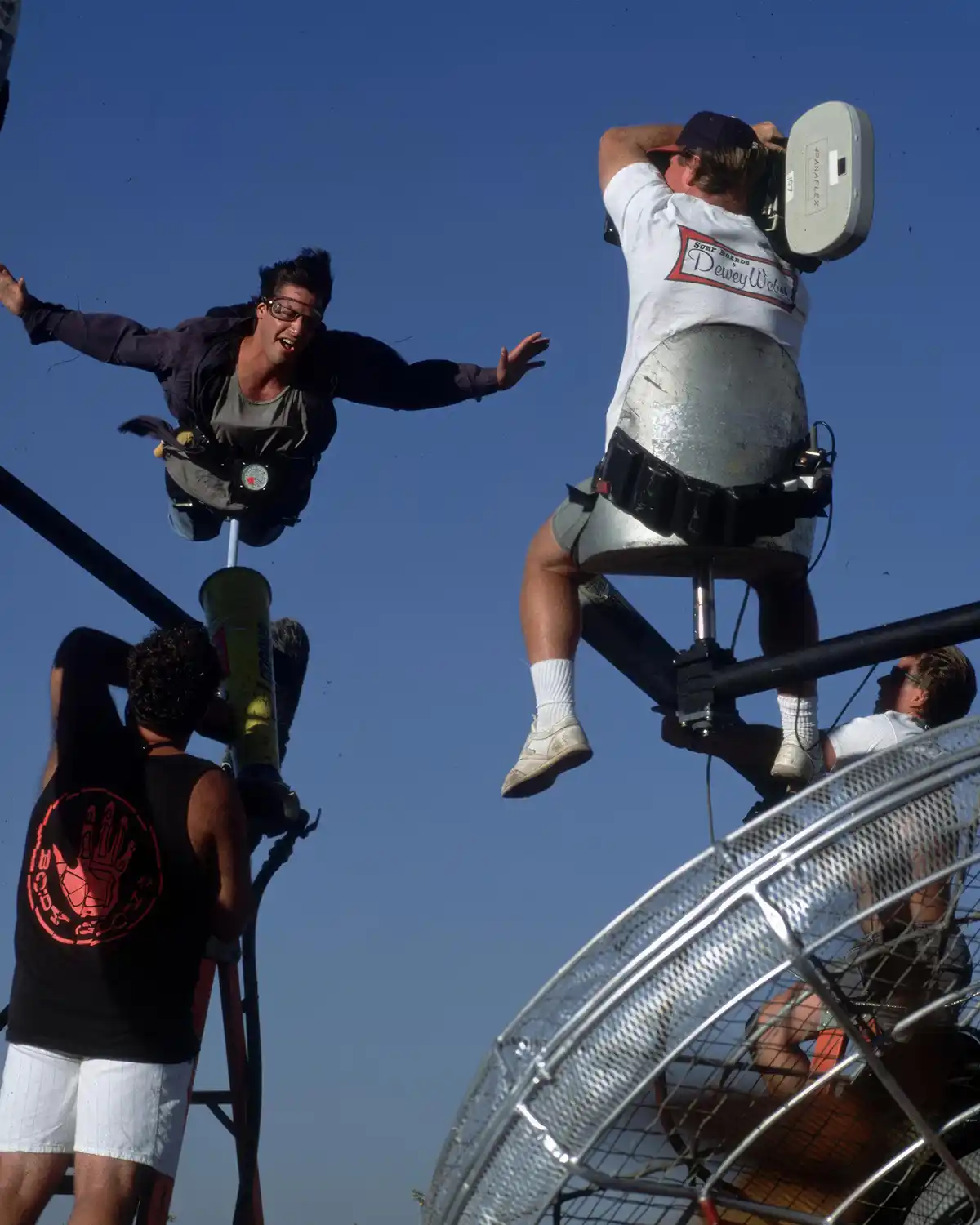 Two men are suspended in mid-air on mechanical platforms against a clear blue sky. The man on the left, wearing glasses and…