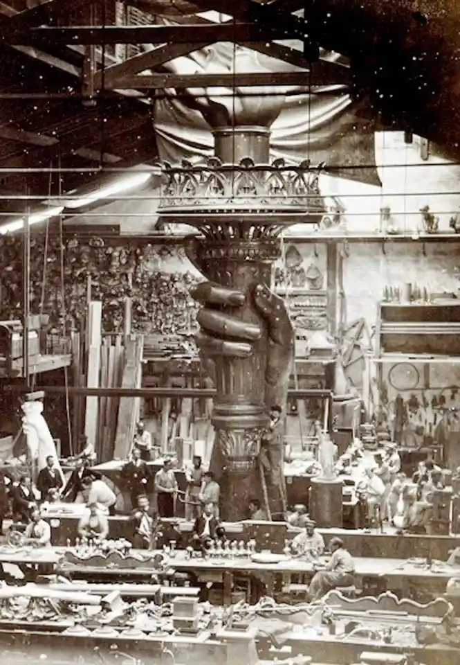 A sepia-toned historical photograph shows the massive torch of the Statue of Liberty being held aloft by a giant sculpted…