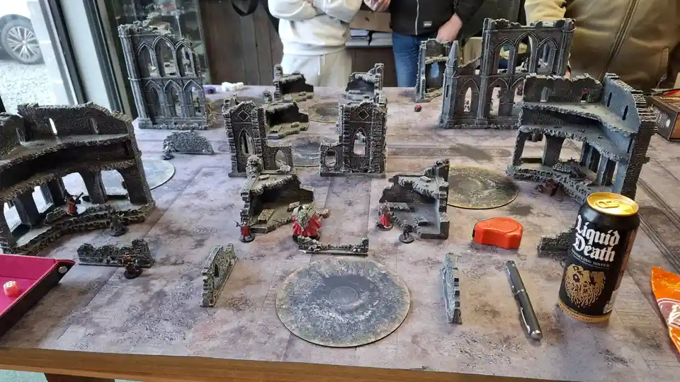 A tabletop wargaming setup featuring miniature figures and ruined castle terrain pieces on a printed battlefield mat. A…