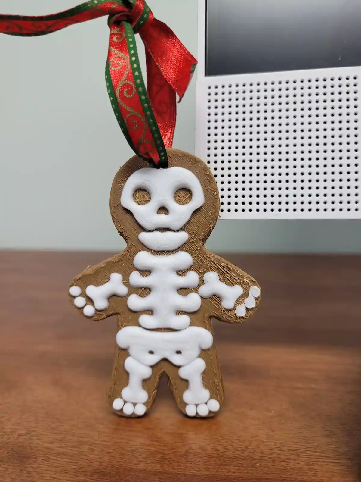 A 3D-printed gingerbread man ornament with a white skeleton design on its brown body, hanging from a red and green ribbon…