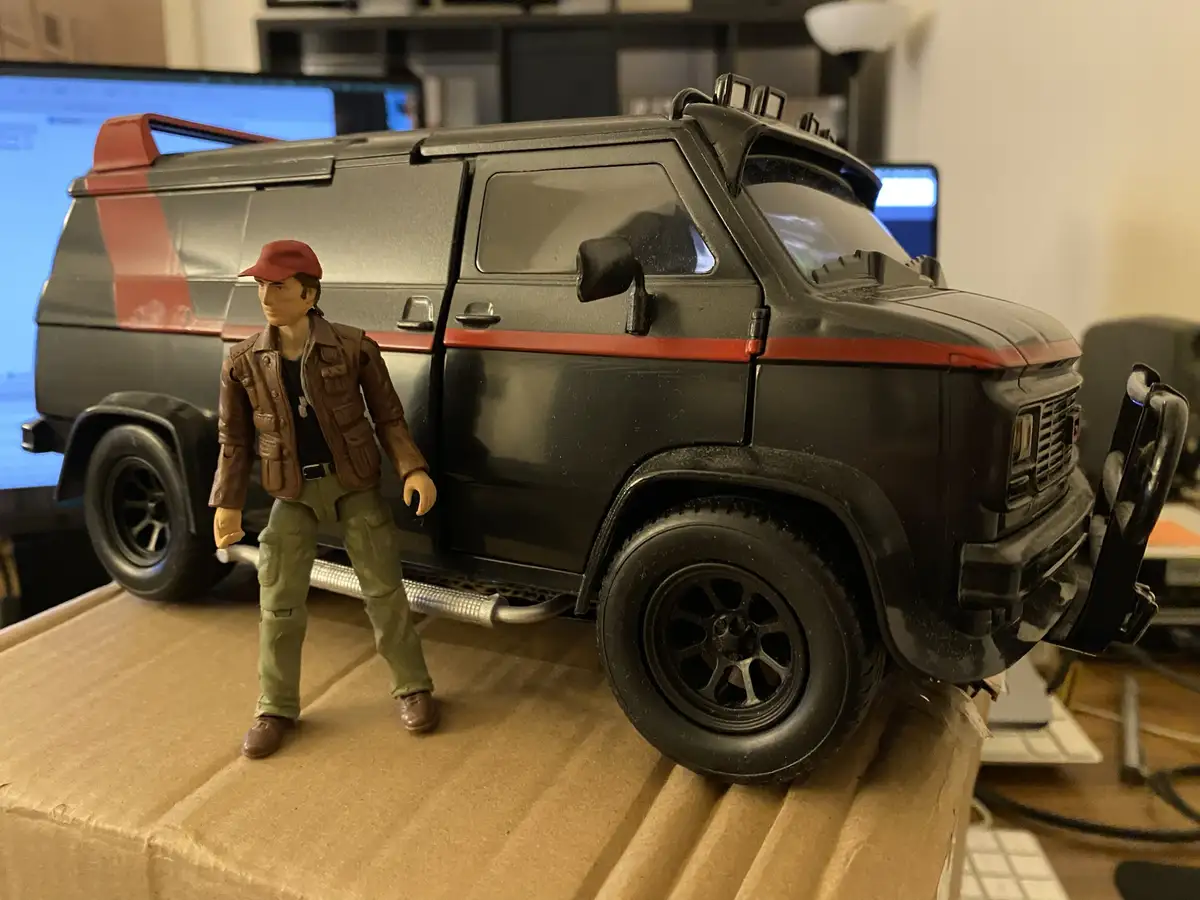 A toy action figure of a man in a red cap, brown jacket, and green pants stands beside a large black toy van with red…