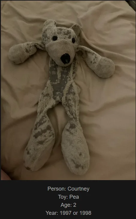 A heavily worn, dirty, and faded stuffed animal resembling a bear lies flat on a beige bedsheet. The toy has black button…