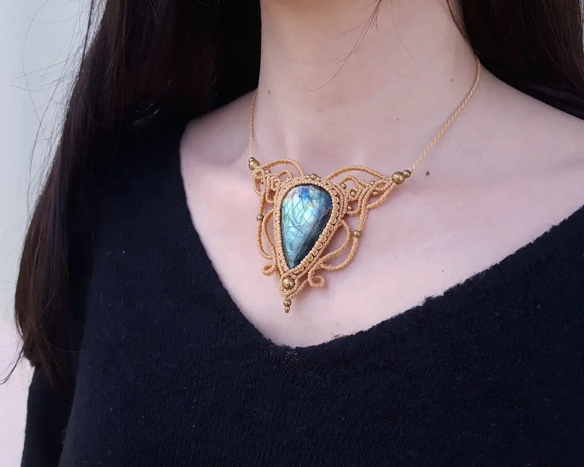 A person with dark hair wearing a black v-neck sweater displays a large, ornate necklace featuring a teardrop-shaped…