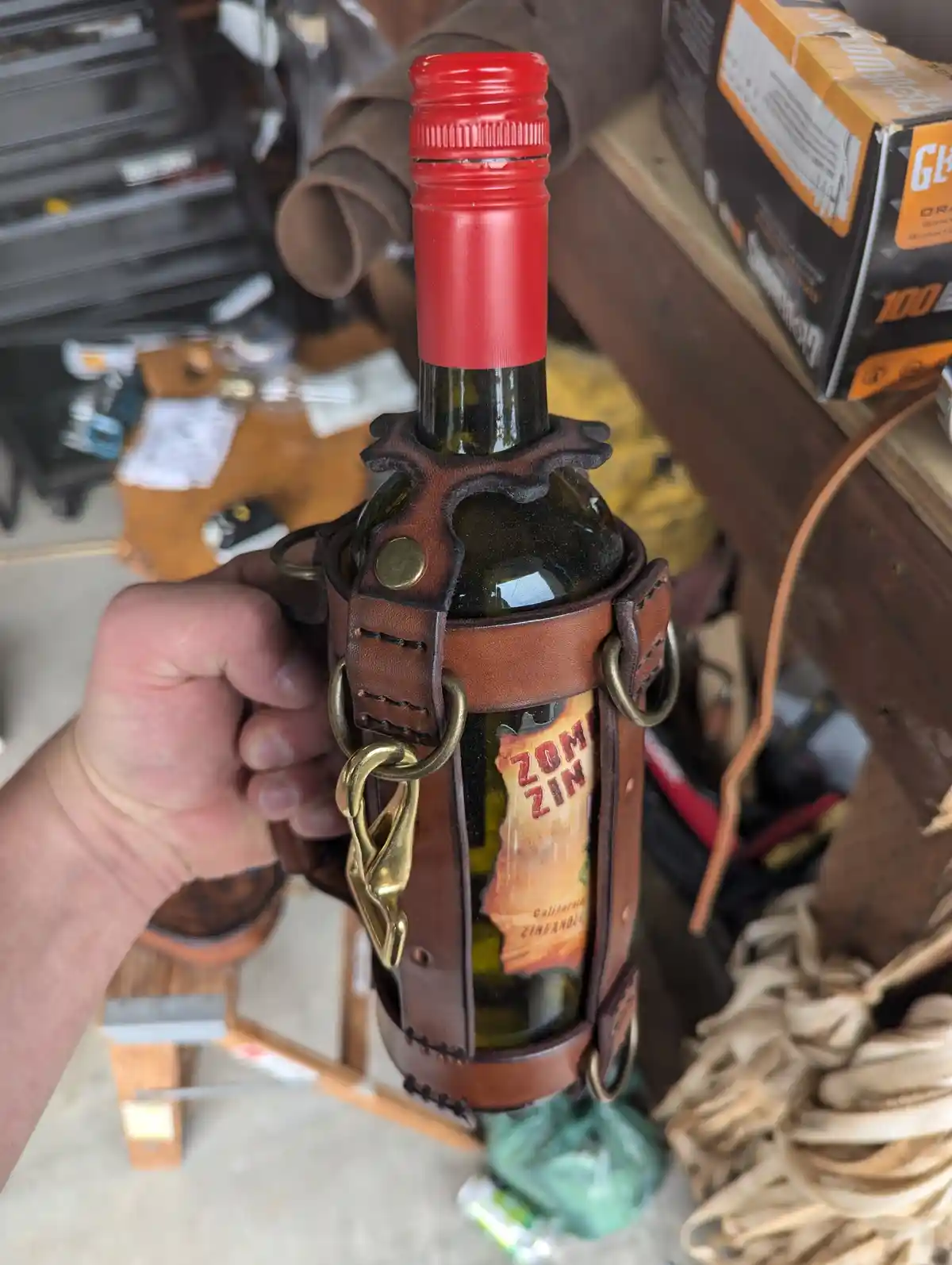 A hand holds a wine bottle labeled 'Zom Zin' encased in a brown leather holster with brass rings and a hook, resembling a…