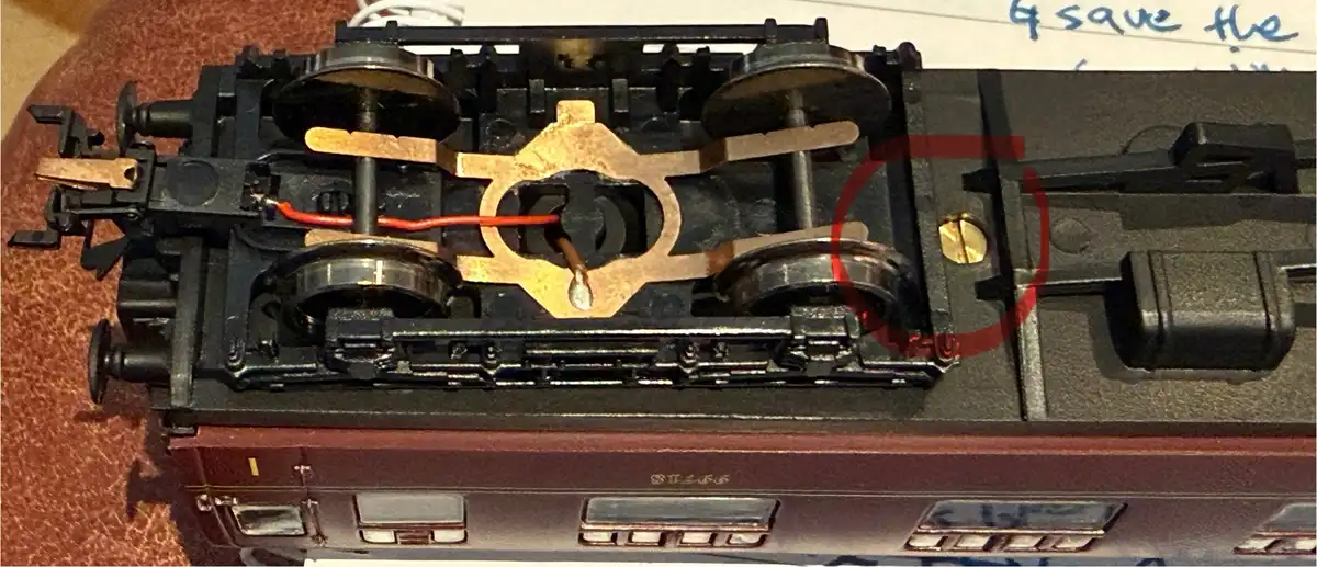 A close-up photo of the underside of a model train, showing its wheels, axles, and electrical contacts. A red circle…