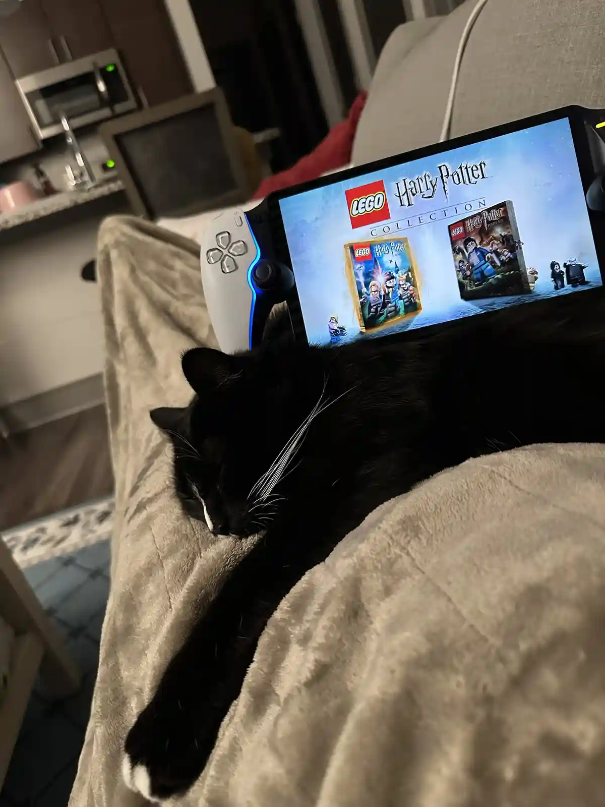 A black cat with white paws is sleeping soundly on a beige blanket, with a PlayStation 5 controller and its screen…