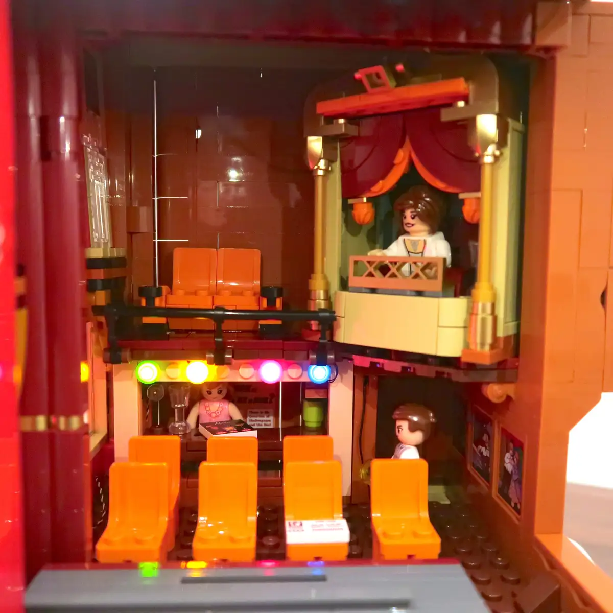 A LEGO theater set with a stage, red curtains, and orange seats. A LEGO minifigure sits on the stage, while another stands…