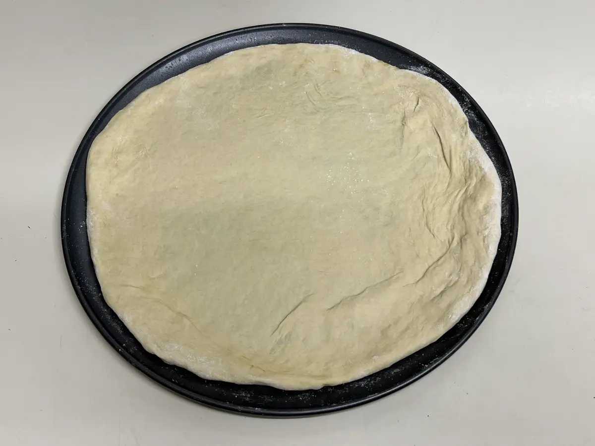 A top-down photo shows a round, pale dough stretched thinly across a black circular pizza pan, with flour dusting the edges…