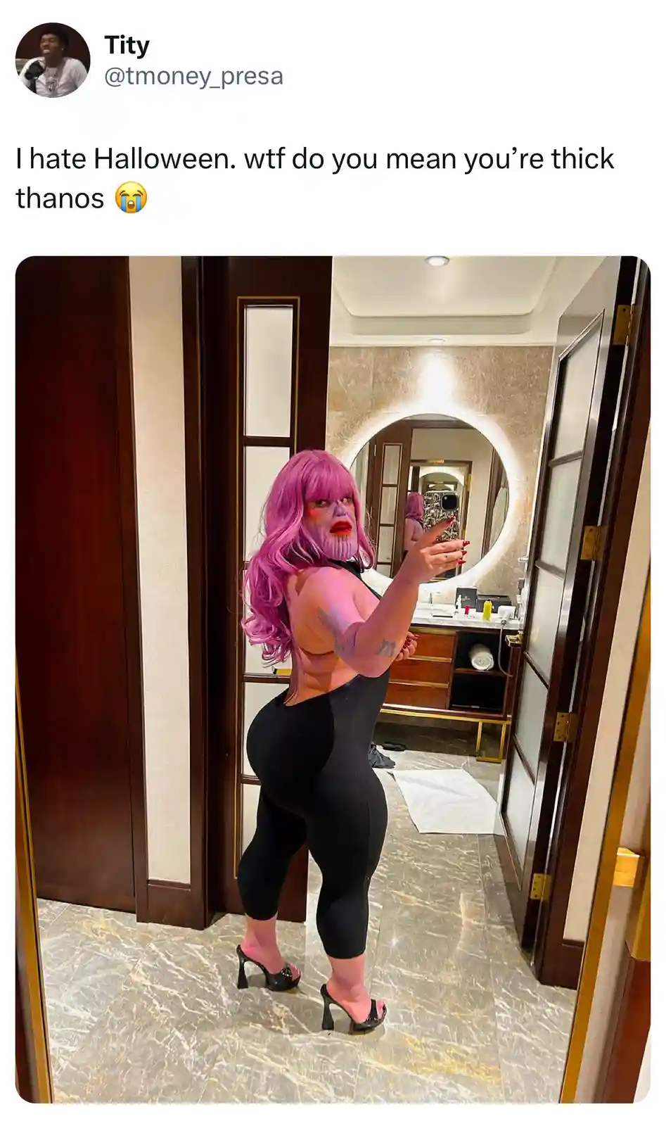 A person with a pink wig and heavy makeup resembling Thanos stands in a luxurious bathroom, wearing a black bodysuit and…
