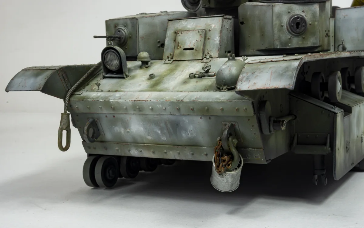 A close-up photo of a detailed, weathered green tank model, showing its tracks, hull, and a bucket hanging from a hook on…