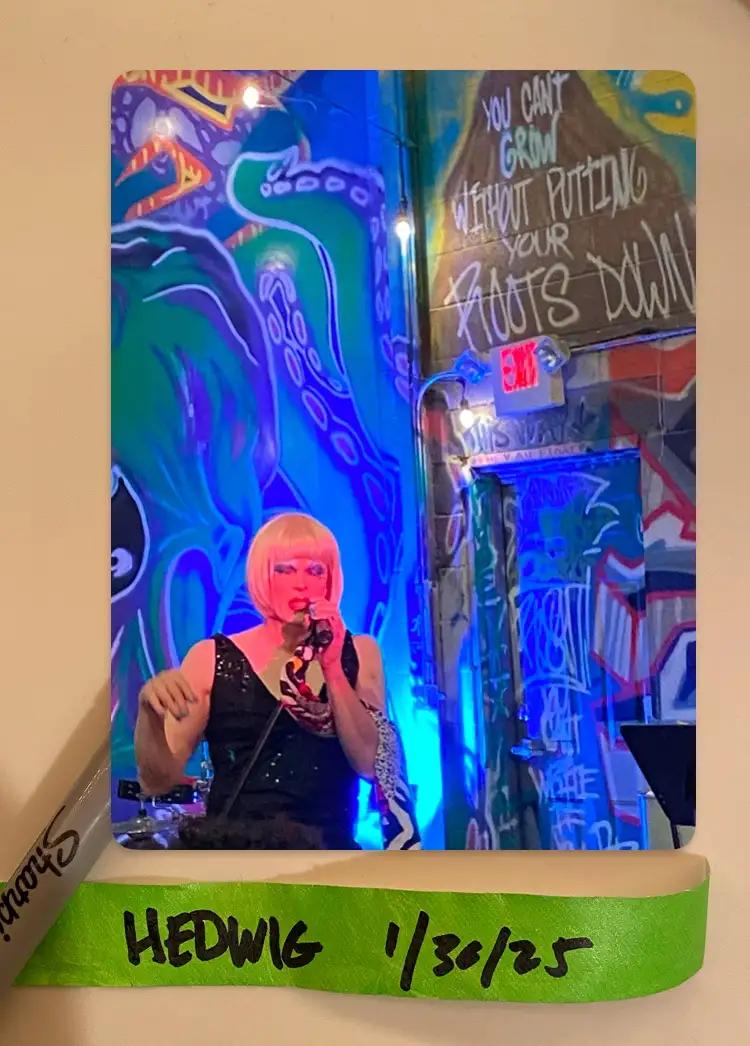 A photo of a performer with a blonde bob wig and red lipstick singing into a microphone on a stage with colorful graffiti…