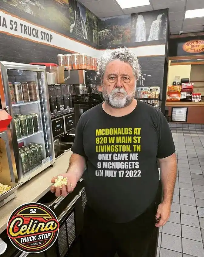 A man with gray hair and a beard stands inside a truck stop, holding a handful of popcorn. He wears a black t-shirt with…