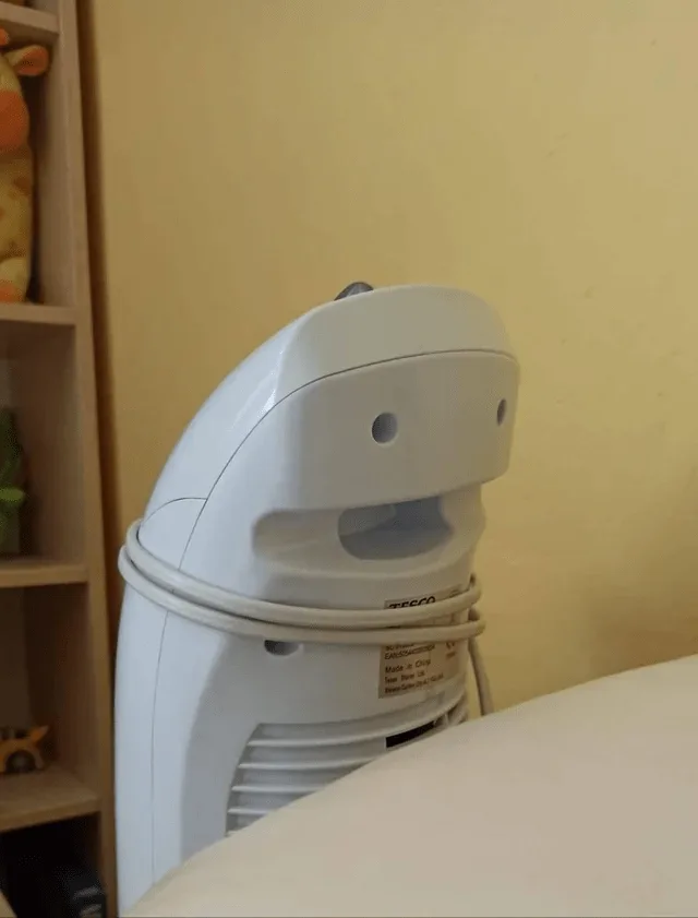 A white, rounded appliance, likely a space heater, is positioned against a yellow wall with a wooden shelf to its left. The…