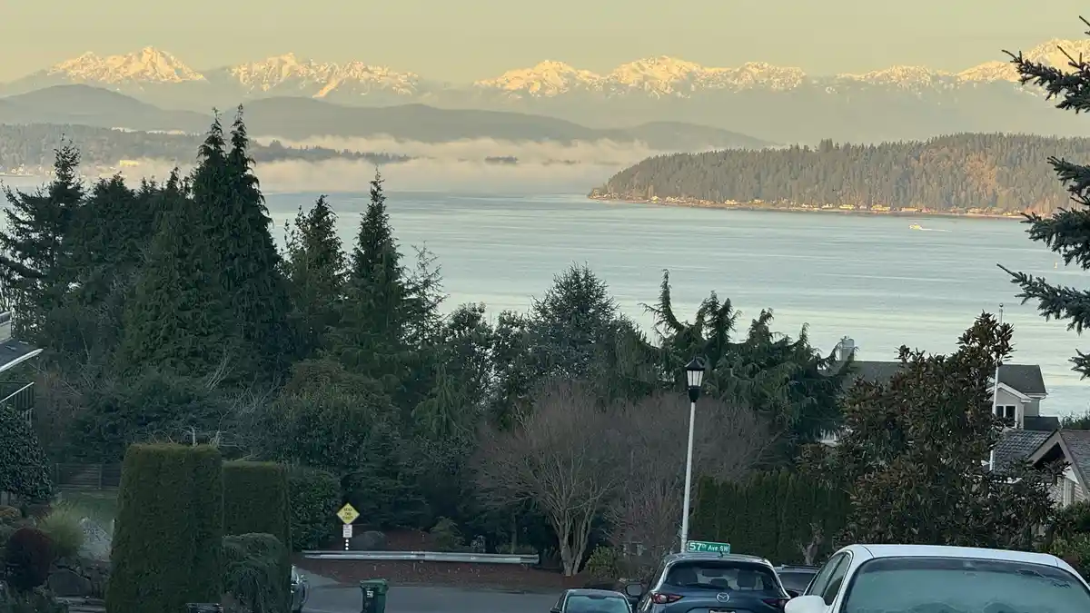 A scenic residential view from a street corner showing snow-capped mountains in the distance, a body of water with mist…