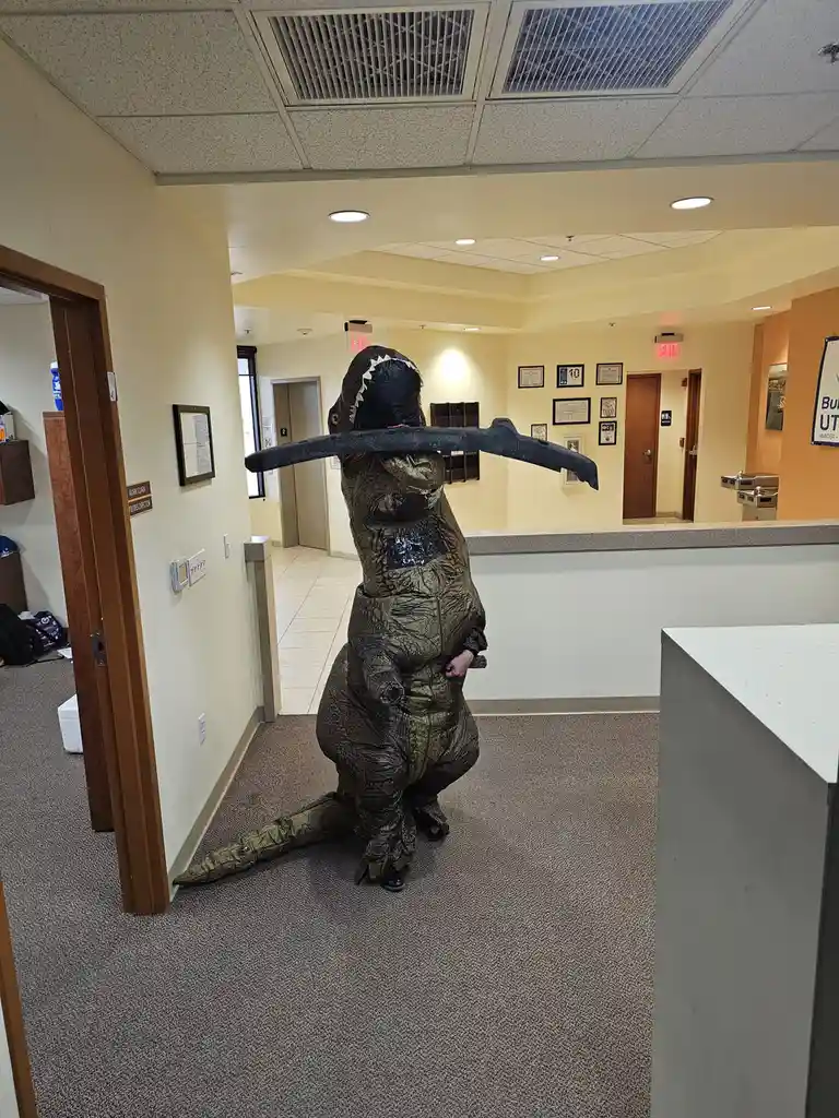 A person in a full-body, green, textured T-Rex costume stands in an office hallway, balancing a large, black, curved scythe…