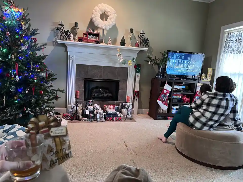 A cozy, festively decorated living room with a lit Christmas tree, fireplace, and presents, where two people sit watching…