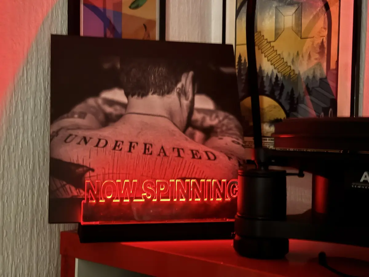 A vinyl record player with a red neon sign reading 'NOW SPINNING' is positioned in front of a framed photo of a man with…
