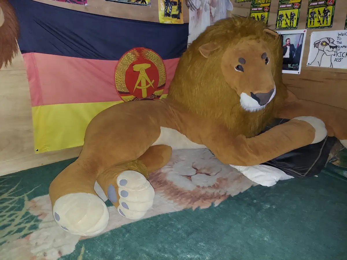 A giant plush lion lies on a rug in a room decorated with a German flag featuring a hammer and compass emblem, action…