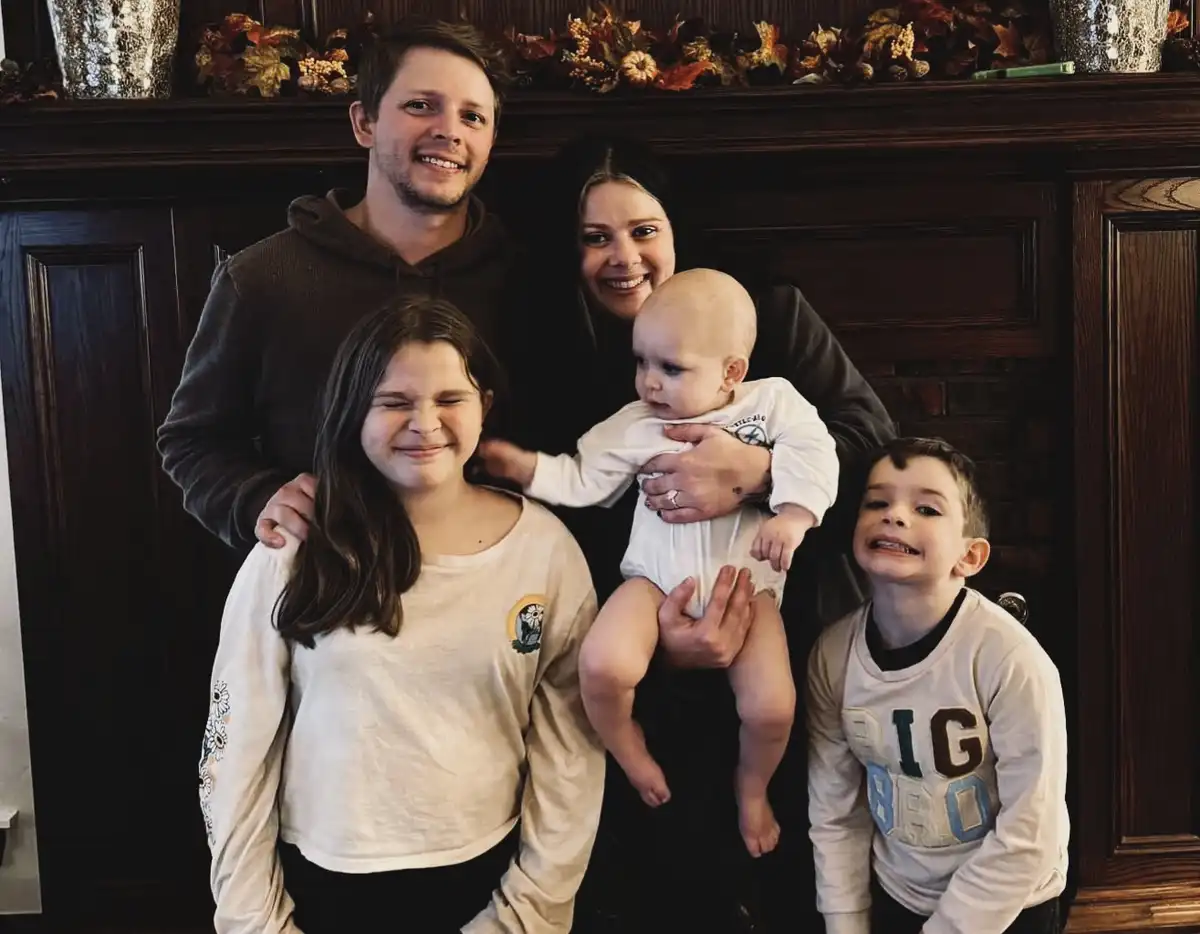 A family photo of five people — a man, woman, baby, and two children — smiling in front of a fireplace. The girl in the…