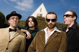 Four men in vintage-style clothing stand outdoors with a distinctive triangular building behind them. The man on the far…
