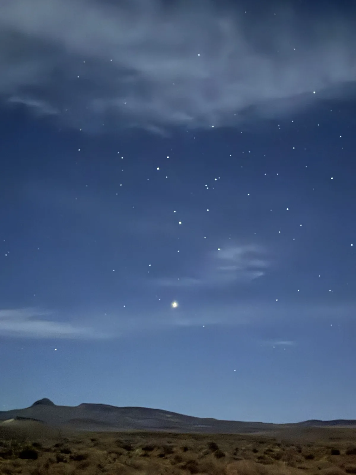 A night sky photo shows a starry expanse with scattered stars and faint clouds above a dark, rolling desert landscape with…