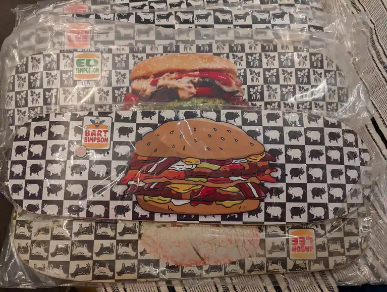 Three skateboard decks in plastic packaging are stacked, each featuring a different burger-themed design on a…