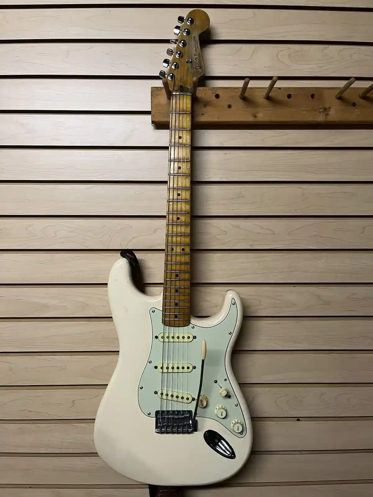 A white Fender Stratocaster electric guitar hangs vertically on a wooden wall-mounted rack against a light wood slat wall…