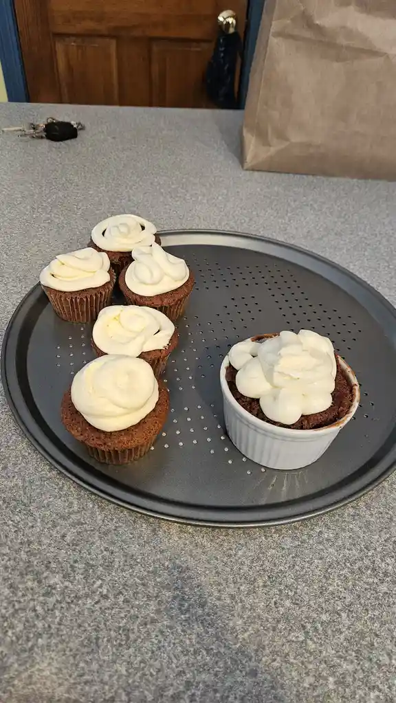 Five cupcakes with neatly piped white frosting sit on a perforated baking tray, while one cupcake in a white ramekin has a…