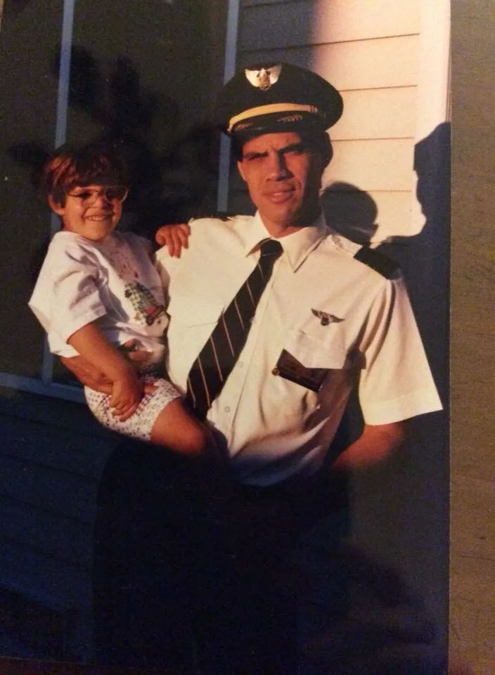 A vintage photo shows a young child with glasses being held by a man in a pilot's uniform, both smiling. The man wears a cap…