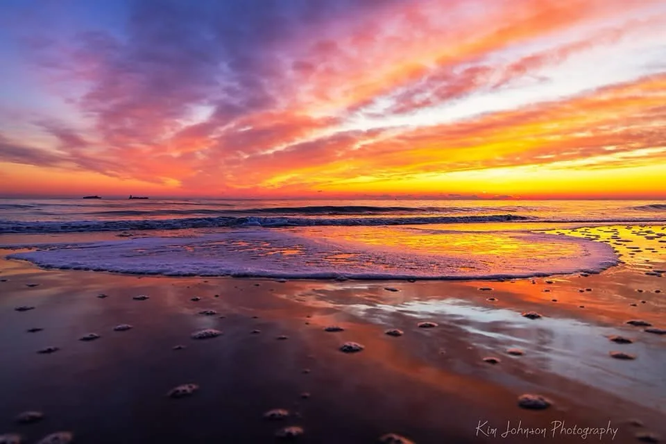 A stunning photograph of a beach at sunset, with vibrant orange, pink, and purple clouds reflecting on wet sand and gentle…