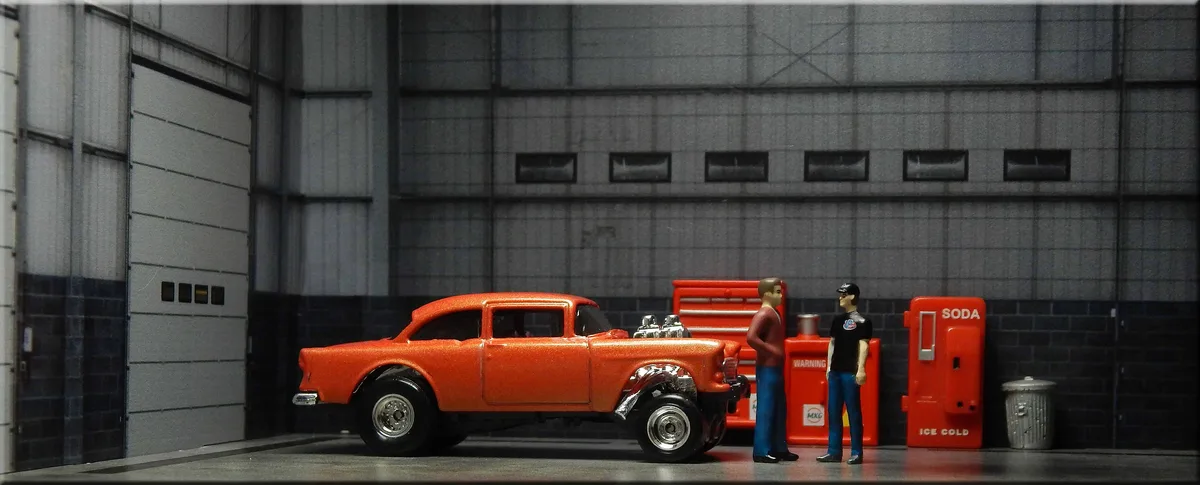 A miniature orange classic car with chrome details and exposed engine sits in a garage diorama next to two tiny figurines…