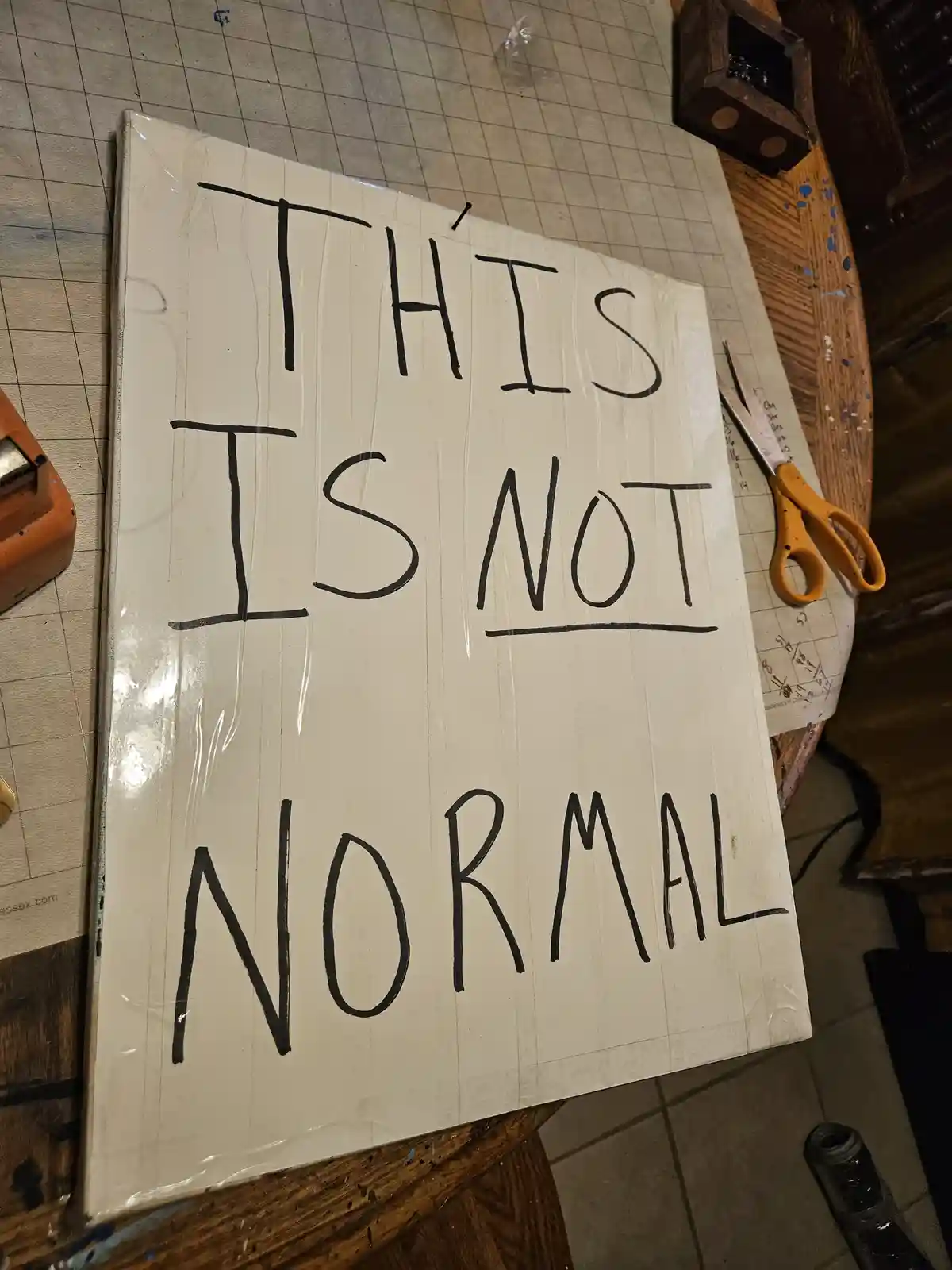 A handmade sign with bold, black handwritten text reading 'THIS IS NOT NORMAL' is taped to a white surface on a cluttered…