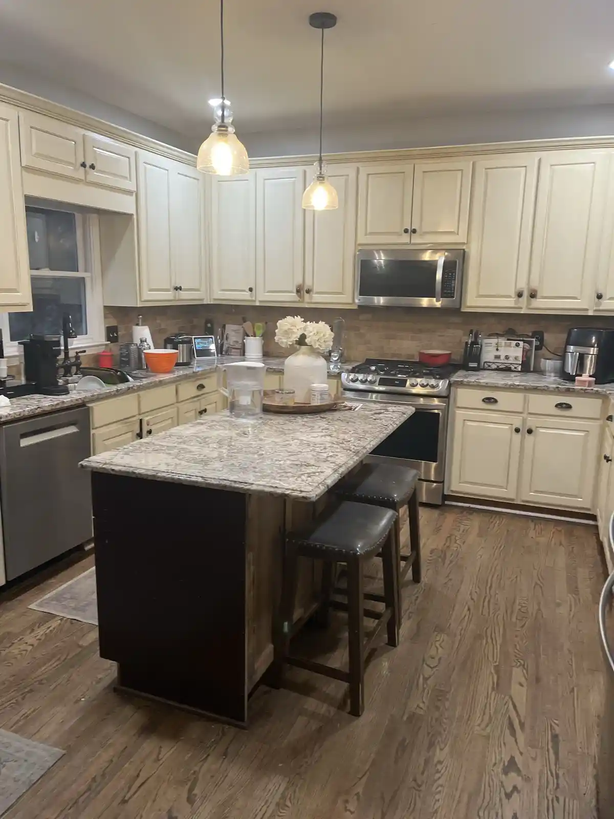 A well-lit, spacious kitchen with cream-colored cabinets, granite countertops, stainless steel appliances, and a central…