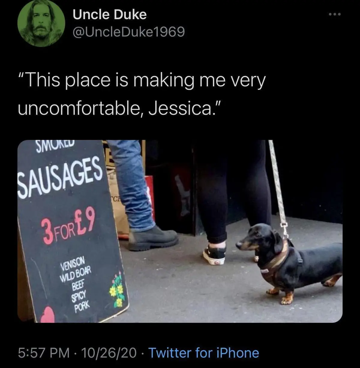 A black-and-tan dachshund on a leash stands near a chalkboard sign advertising 'SMOKED SAUSAGES 3 FOR £9' with meat types…
