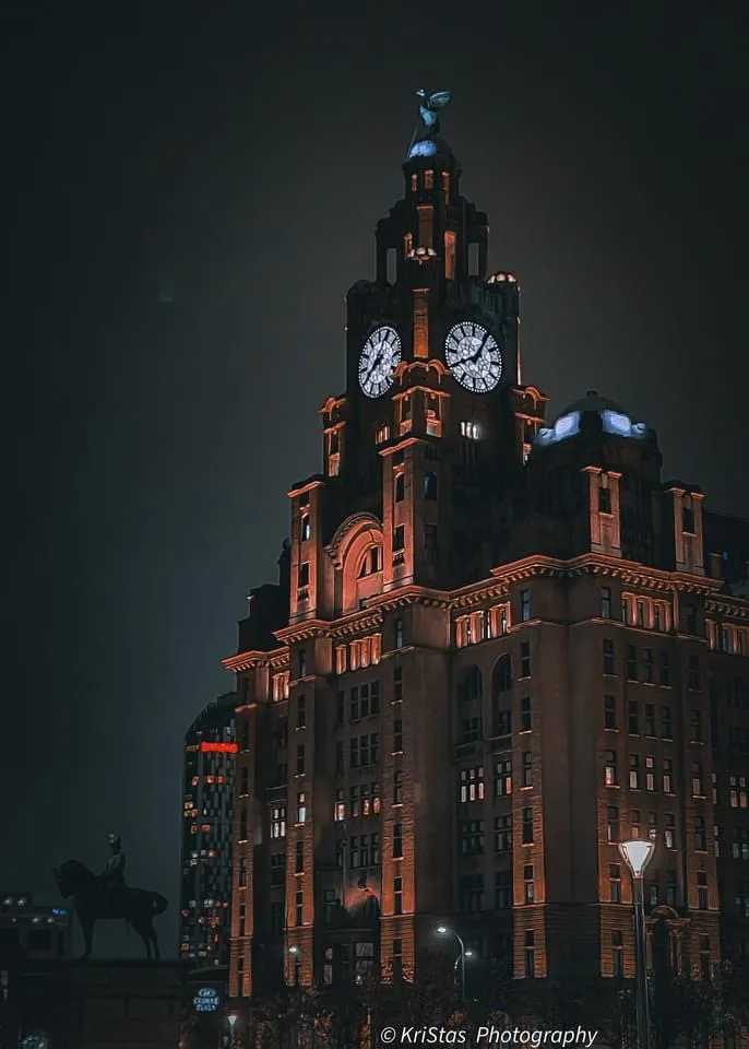 a nighttime photograph of the iconic liver building in liverpool, uk, illuminated with warm orange lights against a dark…