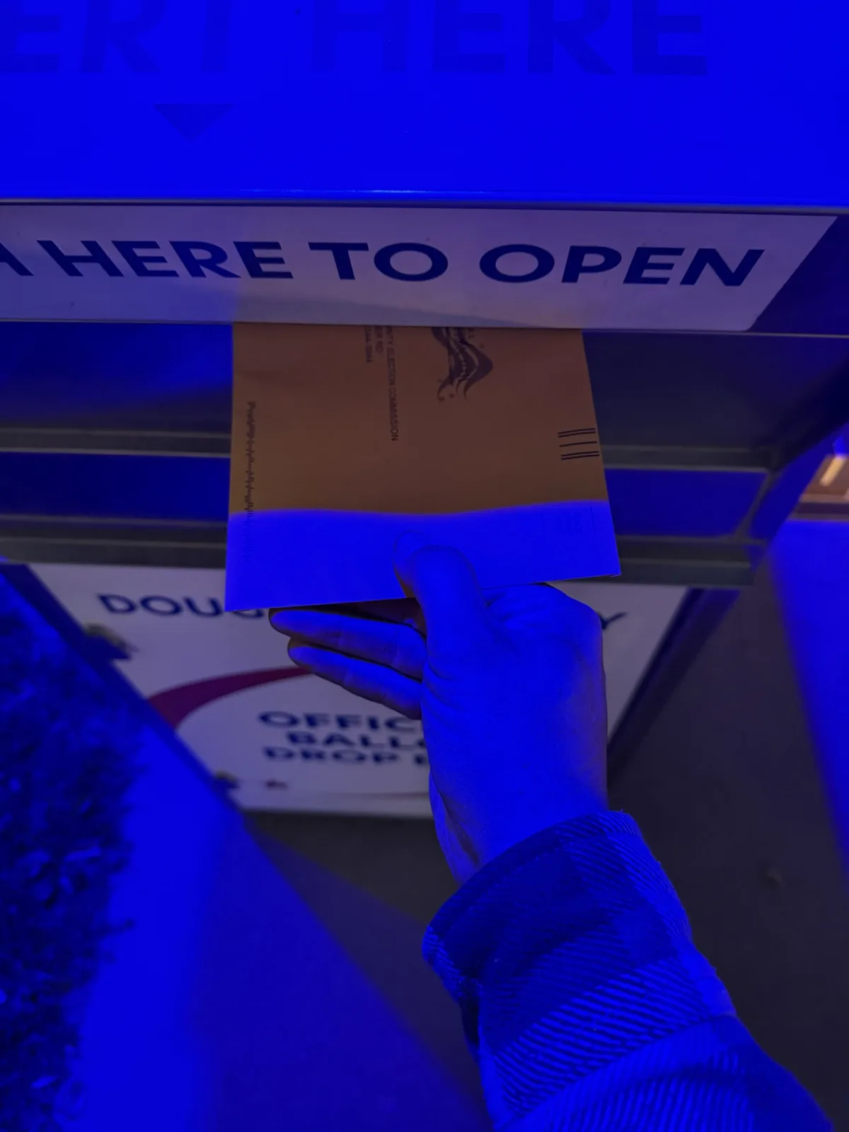 A hand holds a brown ballot envelope in front of a blue ballot drop box with the text 'INSERT HERE TO OPEN' visible above…
